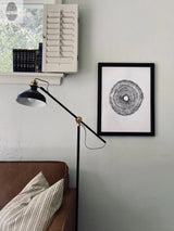 Black and white circular tree ring print framed on gray wall above brown leather chair with patterned pillow, next to black adjustable floor lamp and white shuttered shelf with books.