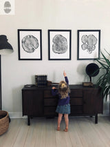 Three black-framed tree block woodcut prints with concentric rings arranged in a row above a dark wooden sideboard, with a young child reaching towards the center print.