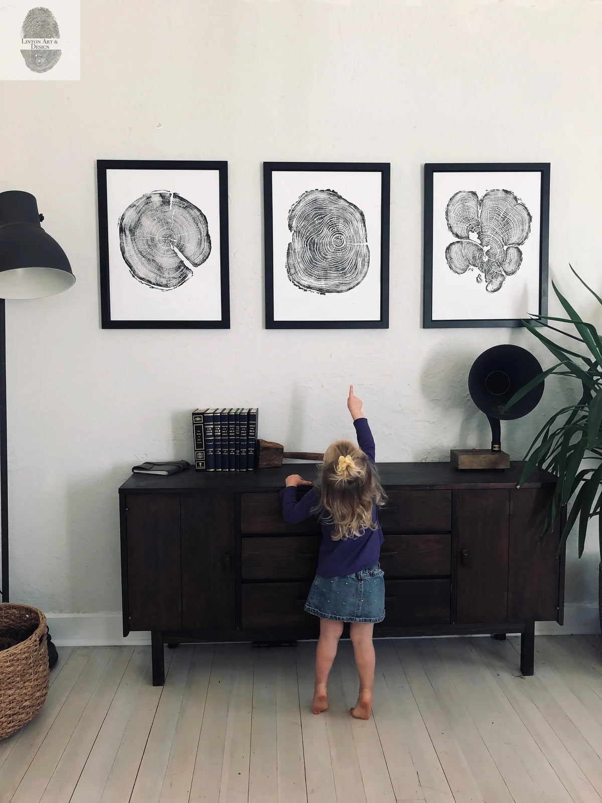 Three black-framed tree block woodcut prints with concentric rings arranged in a row above a dark wooden sideboard, with a young child reaching towards the center print.