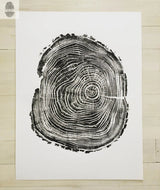 Black woodcut print of a tree cross-section with detailed growth rings and rough bark edges on white paper, centered on a light wooden floor.
