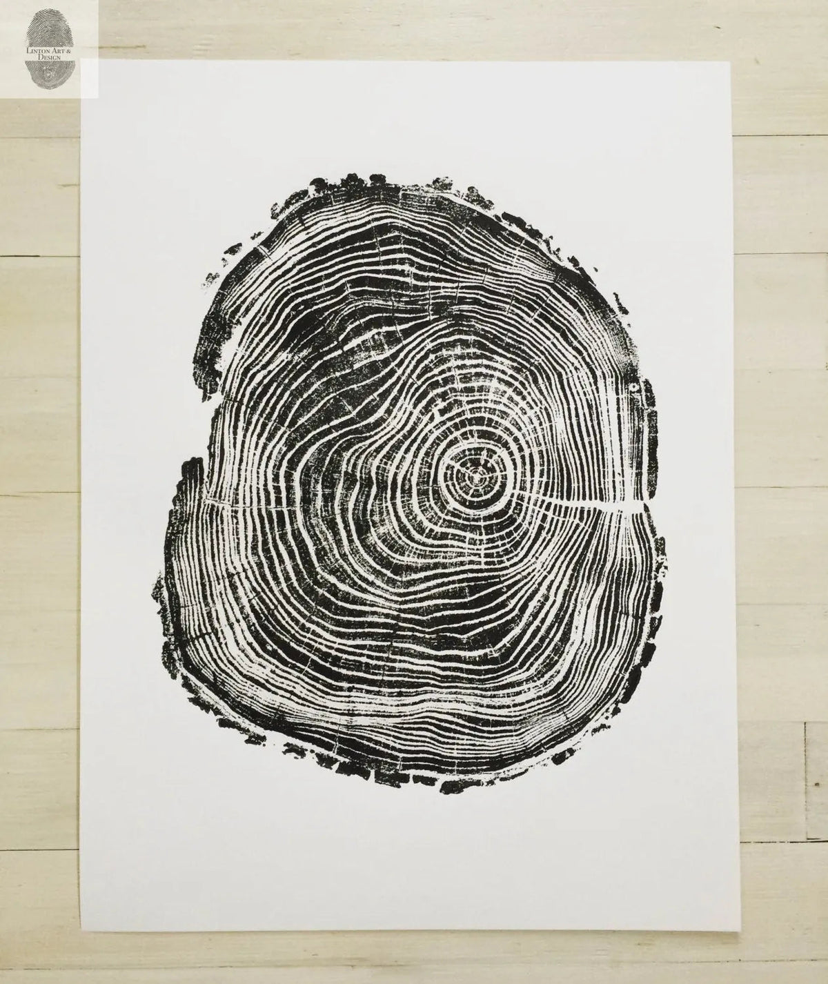 Black woodcut print of a tree cross-section with detailed growth rings and rough bark edges on white paper, centered on a light wooden floor.
