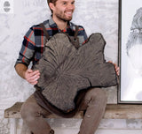 Man in a plaid shirt and apron holding a large dark wood slab with prominent growth rings and an irregular shape, sitting on a rustic wooden bench.