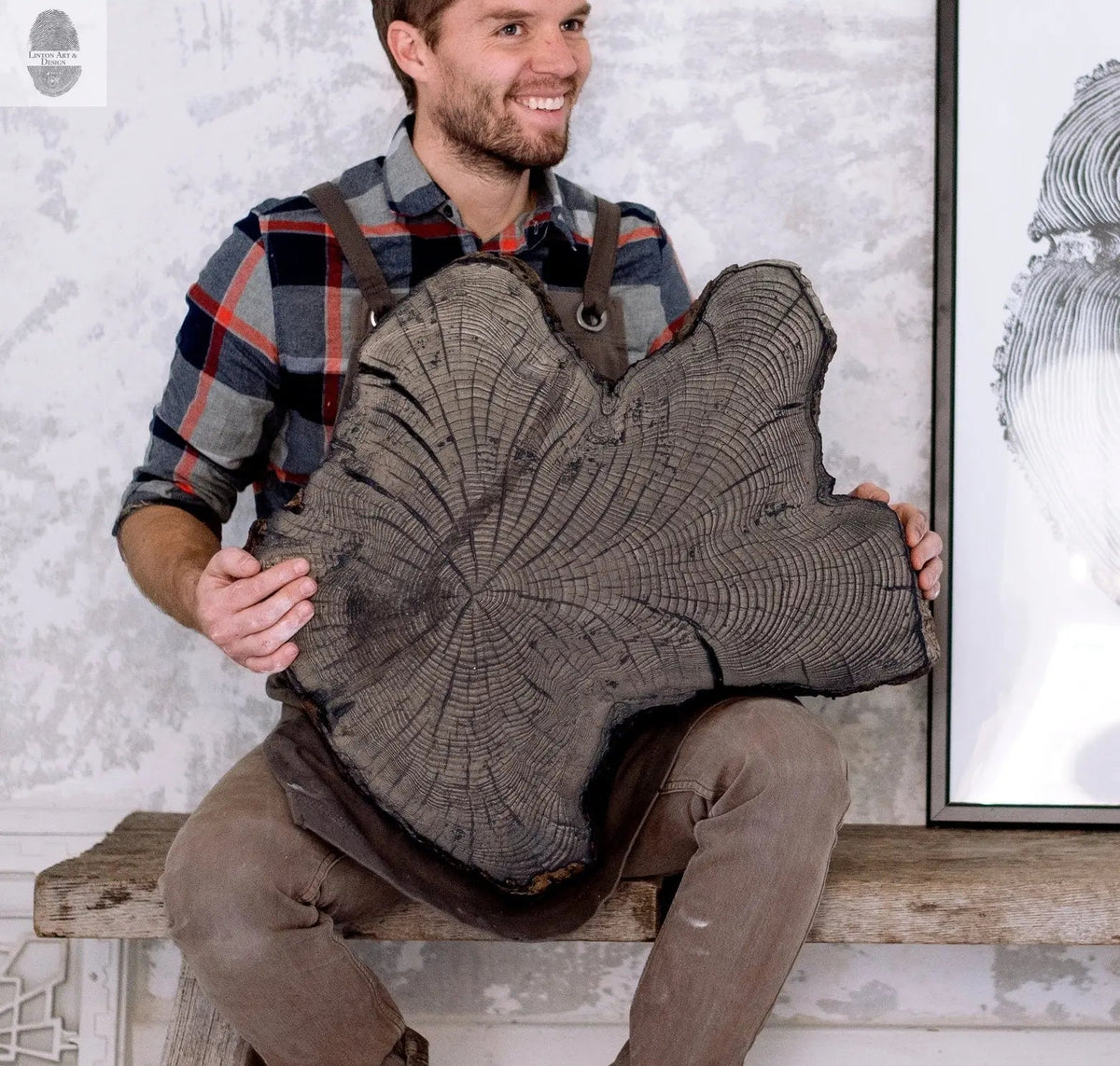 Man in a plaid shirt and apron holding a large dark wood slab with prominent growth rings and an irregular shape, sitting on a rustic wooden bench.