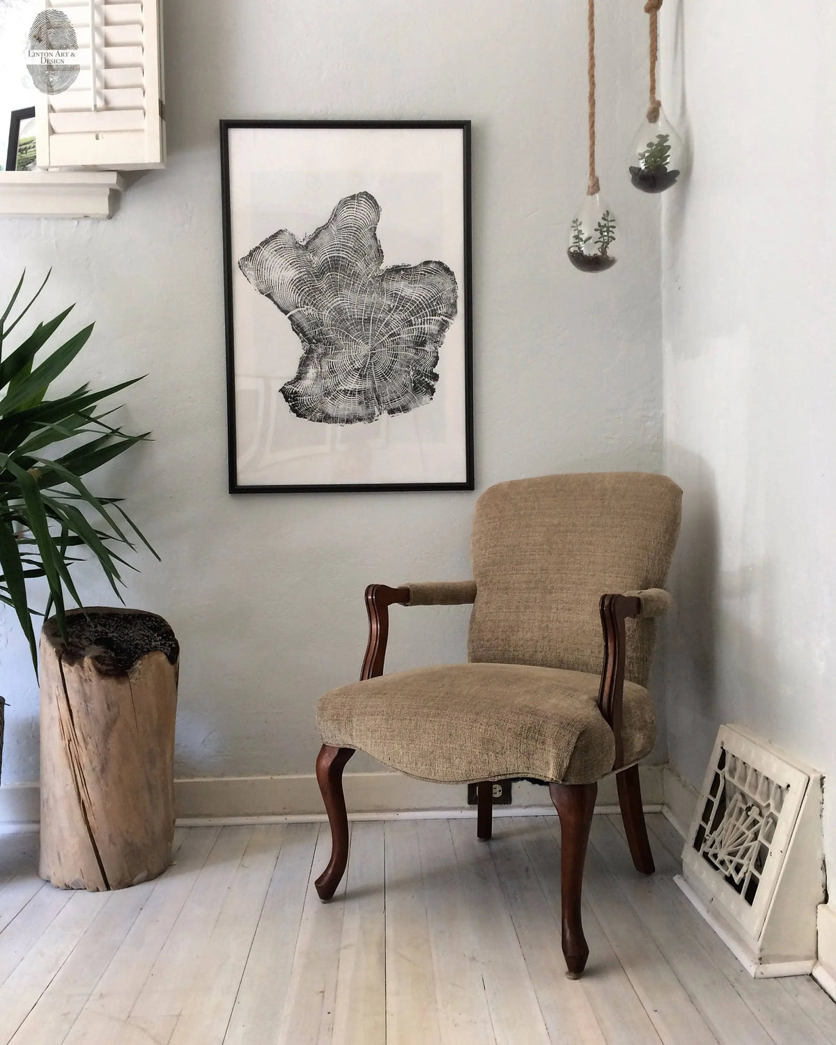 Neutral upholstered wooden armchair with curved legs in a light-walled room featuring black-and-white print of a wood grain pattern and hanging glass terrariums.