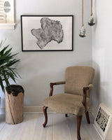 Beige upholstered wooden armchair with carved legs sits next to a tall wood log planter and under a framed print of cross-section driftwood on a white wall.