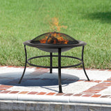 Round black metal fire pit with mesh dome cover and burning firewood, standing on a patterned stone patio with green grass in the background.