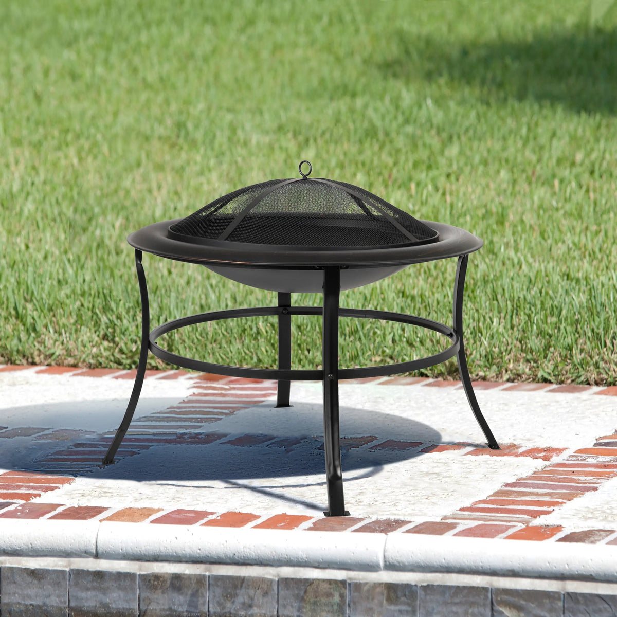 Black round metal fire pit with a mesh dome cover and curved legs, placed on a white and red brick patio with green grass background.