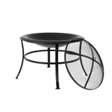 Black round metal fire pit with three curved legs and a removable mesh spark screen leaning against the rim on white background