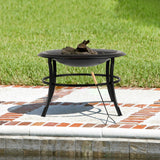 Black metal round fire pit with a wide rim and four legs, holding burnable logs and positioned on a white stone patio with red brick accents near a green lawn.