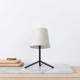 Modern table lamp with a beige linen shade and a tripod black metal base, placed on a marble slab beside stacked light wooden trays with a brass dish.