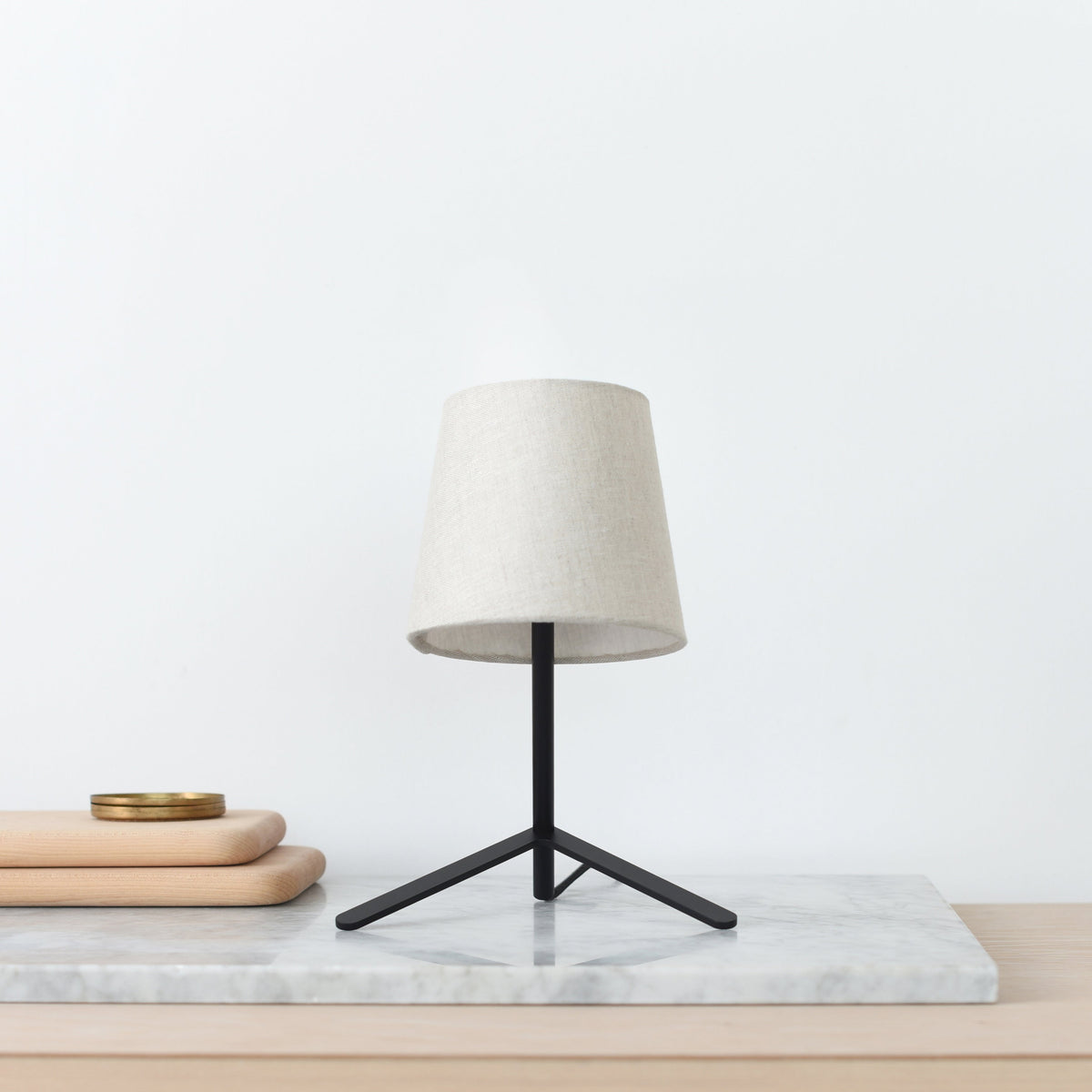 Modern table lamp with a beige linen shade and a tripod black metal base, placed on a marble slab beside stacked light wooden trays with a brass dish.