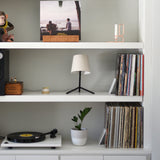 White shelf unit with a small modern black table lamp with beige shade, two rows of vinyl records, a white potted plant, and a turntable on the bottom shelf.