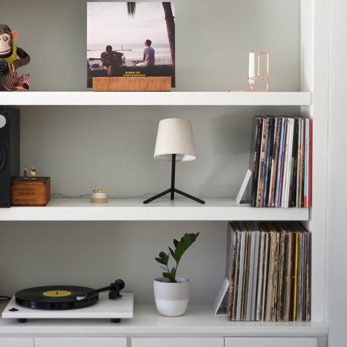 White shelf unit with a small modern black table lamp with beige shade, two rows of vinyl records, a white potted plant, and a turntable on the bottom shelf.