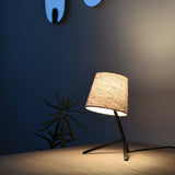 Table lamp with a tilted beige fabric shade on a thin black angular tripod base, casting warm light on a wooden surface against a dim blue wall with abstract art and a plant.