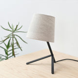 Small table lamp with a tilted off-white fabric shade atop a black three-legged metal base on a light wood surface with a green plant in soft focus behind it