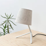 Small table lamp with a tilted fabric drum shade and angled white tripod base, set on a light wood surface with a green plant in the background