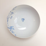 White ceramic bowl with a small blue heart in the center and delicate blue floral patterns along one edge, shown from above on a white background.