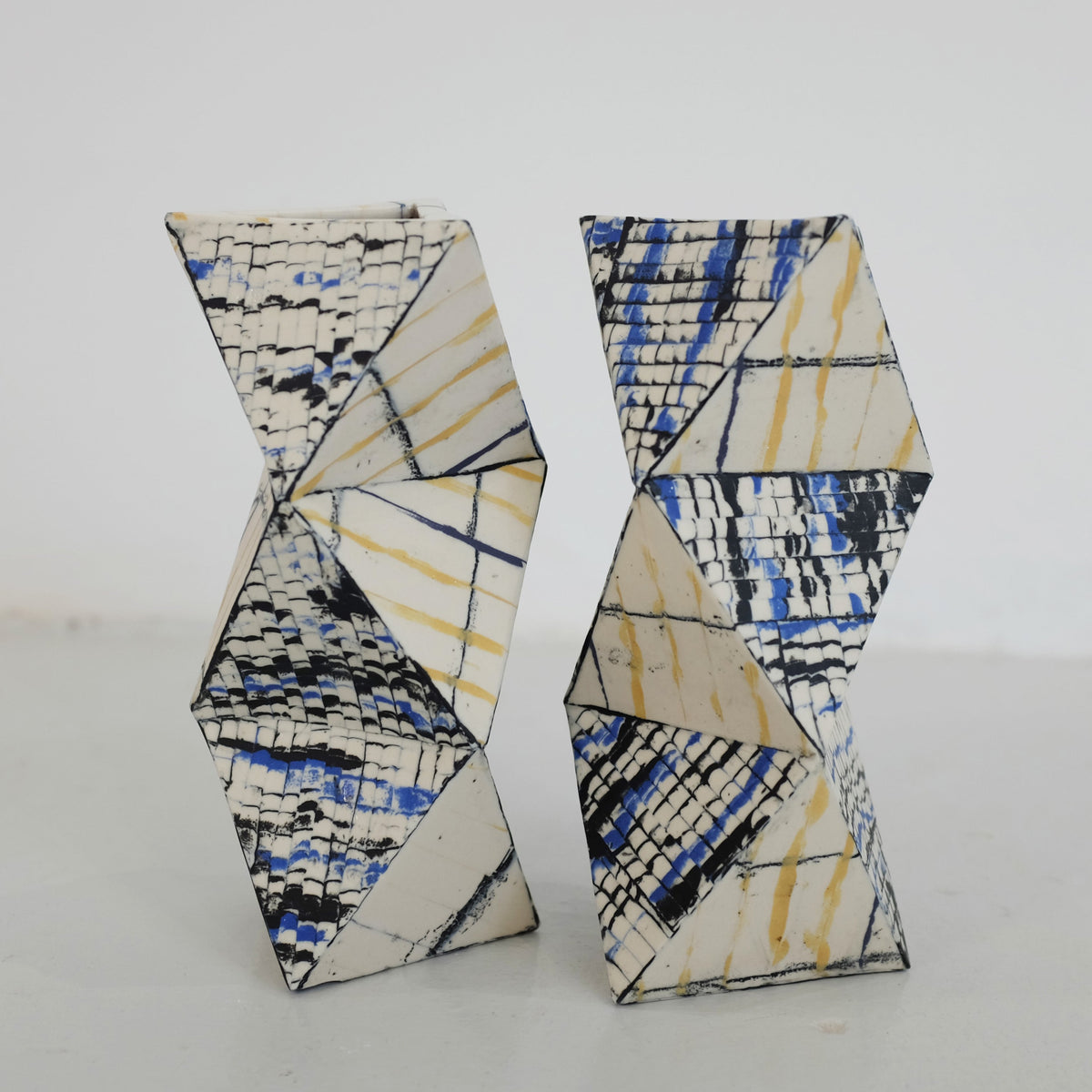 Two angular ceramic vases with zigzag silhouettes, featuring abstract black, blue, and yellow linear patterns on a white background, placed on a plain surface.