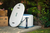 White inflatable tub with textured surface and black handles, partially inflated outdoors next to a R E V X branded pump and cardboard boxes, surrounded by greenery.