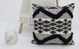 Square decorative pillow with black tufted chevron and diamond patterns on a white woven background, placed on a white surface with a fringed beige throw and small cream ceramic decor nearby.