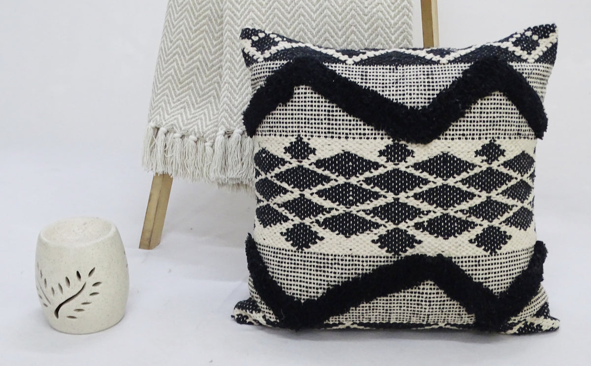 Square decorative pillow with black tufted chevron and diamond patterns on a white woven background, placed on a white surface with a fringed beige throw and small cream ceramic decor nearby.