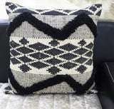 Square pillow with black tufted chevron stripes and diamond patterns on a textured cream background resting on a couch.
