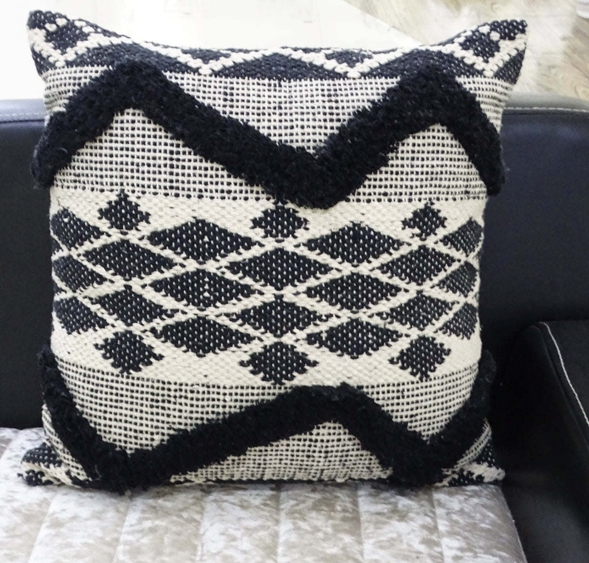 Square pillow with black tufted chevron stripes and diamond patterns on a textured cream background resting on a couch.