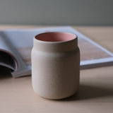 Beige ceramic coffee tumbler with a smooth matte finish and soft pink interior, sitting on a light wood surface in front of an open book with blurred pages.