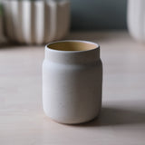 Matte light gray ceramic coffee tumbler with a slightly flared rim, set on a smooth light wood surface with blurred pottery in the background.