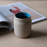Matte beige ceramic coffee tumbler with a smooth blue interior rim, placed on a light wooden surface beside an open magazine with a blurred image of a person in red.