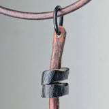 Pendant featuring a small, finished wood piece with a twisted black textured metal band wrapped around its middle, hanging from a leather cord with a metal ring.