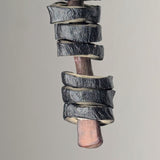 Close-up of a dark, textured spiral wood shaving curling tightly around a reddish-brown pencil with a smooth surface, against a neutral background.