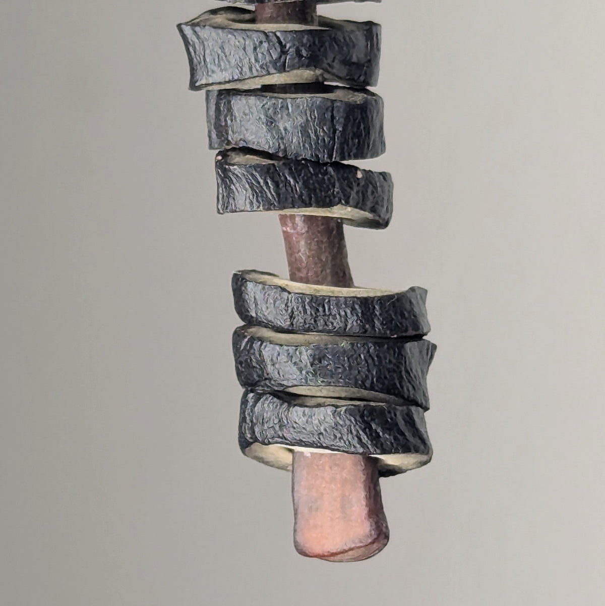Close-up of a dark, textured spiral wood shaving curling tightly around a reddish-brown pencil with a smooth surface, against a neutral background.