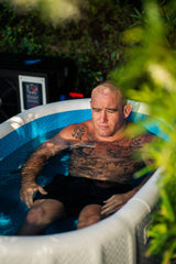 Man with tattoos relaxing in a white and blue inflatable tub filled with water, surrounded by greenery outdoors.