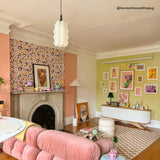 Living room with a curved pink velvet sofa, a patterned wallpaper accent above a stone fireplace, and an arrangement of colorful framed art on a light green wall.