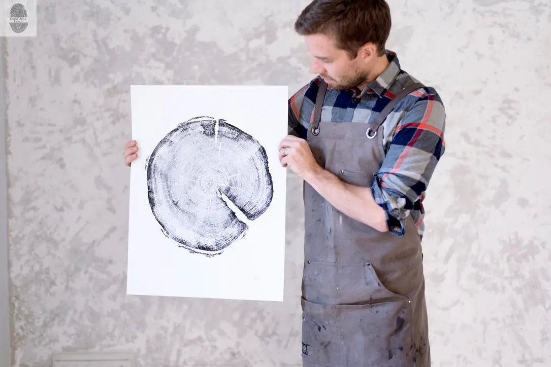Man in gray apron and plaid shirt holding a black and white circular print resembling a tree cross-section against a textured gray wall.