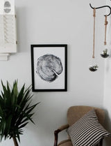 Black and white framed print of a tree ring cross-section hung on a white wall above a beige armchair with a black and white houndstooth pillow, next to green potted plant and hanging glass planters.