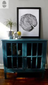 Black and white framed art print of a tree cross-section hung above a teal wooden cabinet with glass doors, featuring a small clock, a potted plant, and a yellow decorative object.