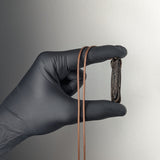 Black-gloved hand holding a dark brown elongated pendant with textured vertical grooves, suspended on a thin brown cord against a plain light gray background.