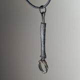 Dark metal pendant shaped like a textured twig with a twisted twig loop at the bottom, hanging from a black leather cord against a neutral background.