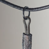 Metal pendant with a textured, tree-bark-like surface and a looped top, hanging from a dark, worn leather cord against a neutral background.