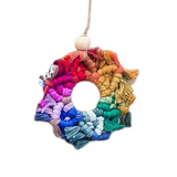 Circular macrame ornament with looped and fringed yarn radiating in a rainbow gradient from red to violet, hanging from a jute string with a wooden bead at the top.