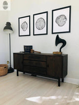 Three black-and-white tree cross-section prints in black frames hang above a dark wooden sideboard topped with vintage books and a black gramophone against a white wall.