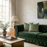 Deep green velvet tufted sofa with two geometric-patterned lettuce green pillows against a textured light gray wall featuring an abstract green and gold painting