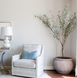 Light gray armchair with a blue and white geometric patterned pillow, placed next to a round side table with a textured white lamp and a large olive tree in a tall pot.