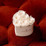 Seven creamy white roses arranged in a small, beige heart-shaped box with 'The Million Roses' logo, placed on rich, textured burnt orange fabric.