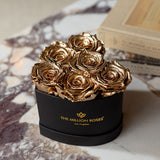 Five metallic gold roses arranged in a black heart-shaped box with gold logo text, placed on a white and brown marbled surface beside books.