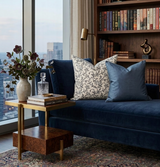 Three decorative pillows on a deep blue velvet sofa, including a cream-colored pillow with indigo vine floral pattern flanked by solid indigo blue pillows, in a cozy reading nook with bookshelves and city view.