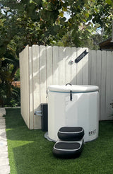 White inflatable barrel hot tub with quilted texture and black inflatable steps, set on green artificial grass near a white wooden fence and leafy tree.
