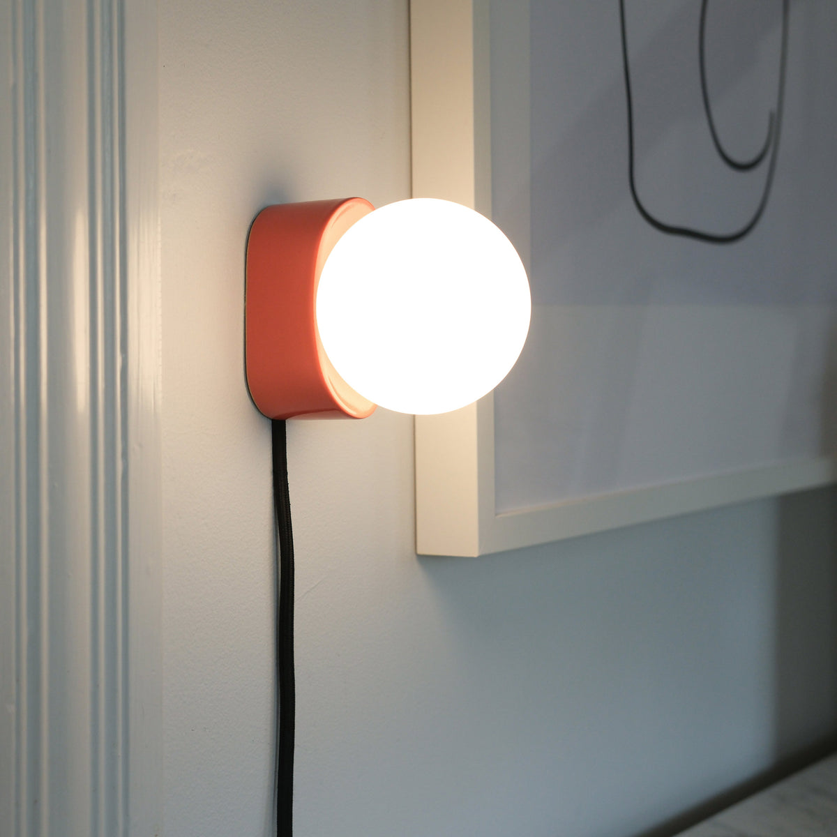 Wall-mounted sconce featuring a glowing white glass orb on a small matte coral pink base with a visible black cord against a pale blue-gray wall.